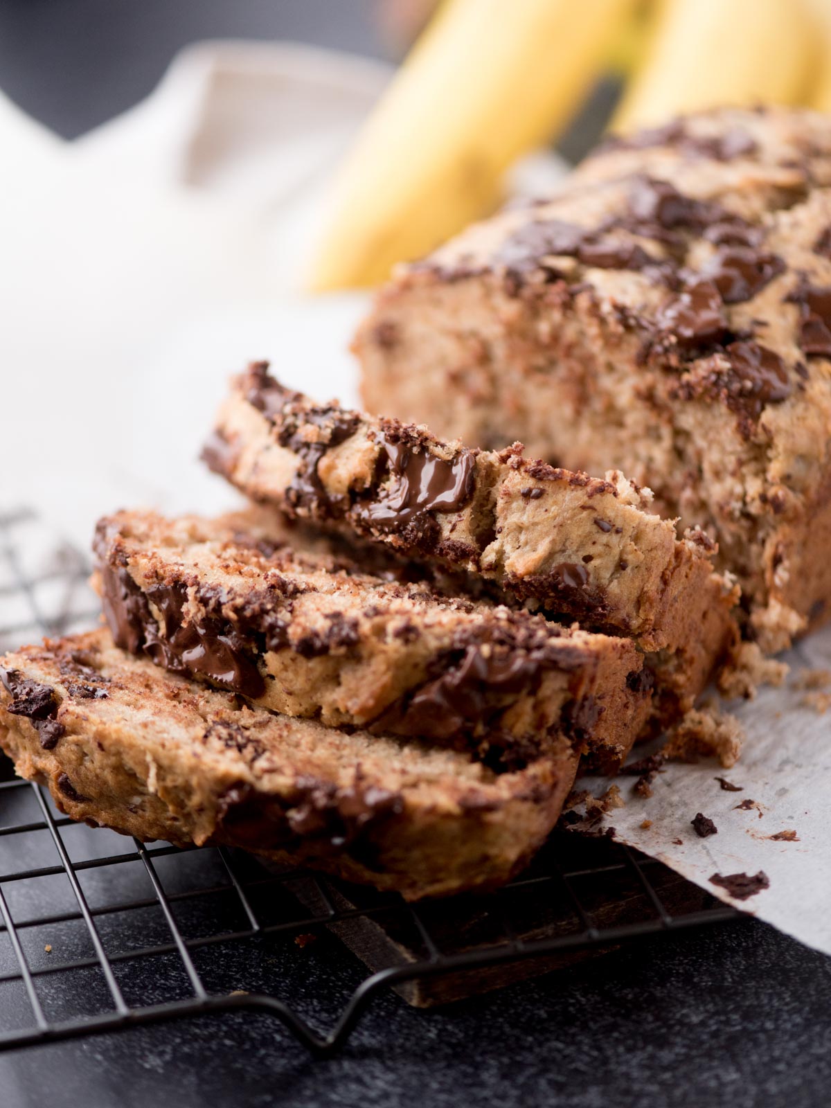 Sliced chocolate chip banana bread on a cooling rack, with melted chocolate chunks visible in the moist bread. Whole bananas are blurred in the background.