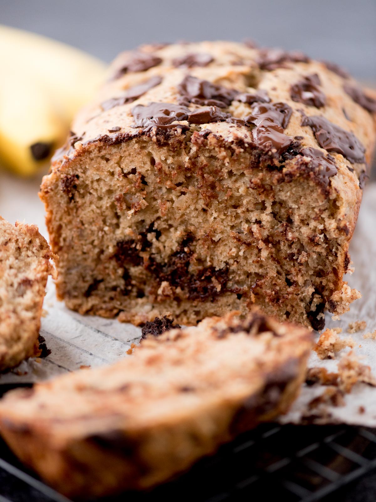A close-up of a sliced loaf of banana bread with visible chocolate chunks, resting on parchment paper. The bread looks moist, with a golden-brown crust and chocolate pieces melted on top. A banana is blurred in the background.