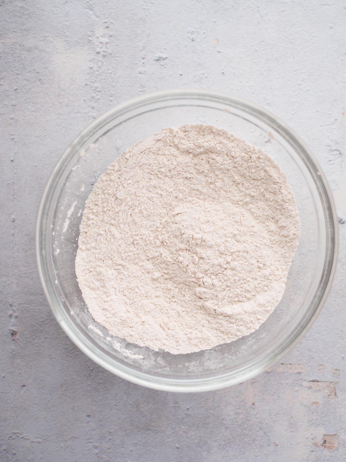 A clear glass bowl filled with a mound of white flour sits on a light gray textured surface.