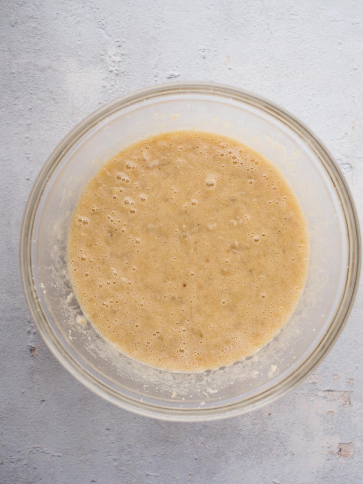 A glass bowl filled with a tan, bubbly batter sits on a light gray textured surface, viewed from above.