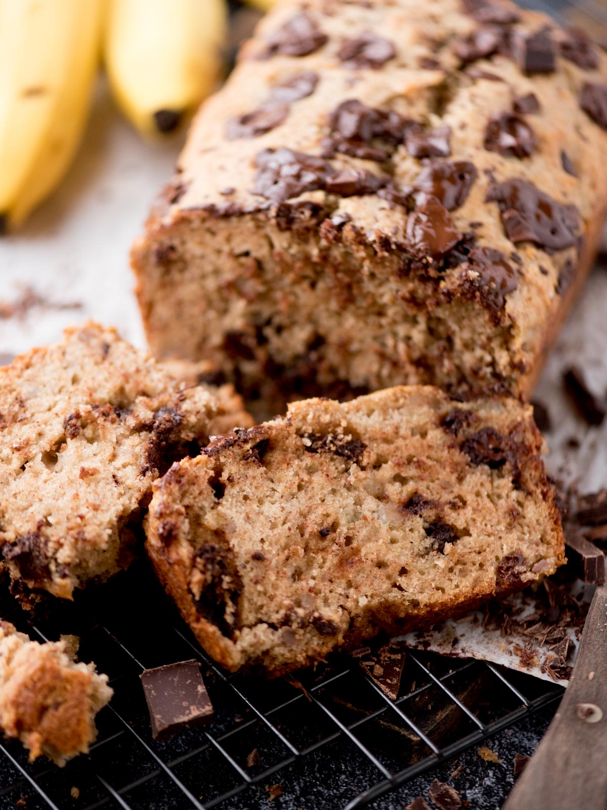 A close-up of sliced chocolate chip banana bread on a cooling rack, with a loaf in the background and bananas partially visible. The bread is moist, dotted with chocolate chunks.