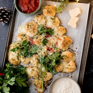 Garlic bread bites arranged in a Christmas tree shape, topped with herbs and cheese, garnished with parsley. Served with red marinara and white dipping sauces on a parchment-lined baking tray.