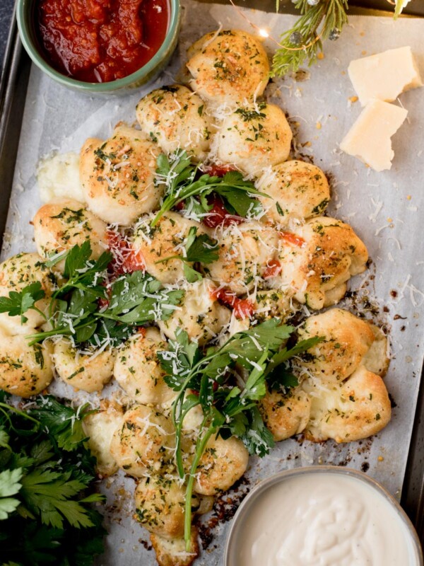 Garlic bread bites arranged in a Christmas tree shape, topped with herbs and cheese, garnished with parsley. Served with red marinara and white dipping sauces on a parchment-lined baking tray.