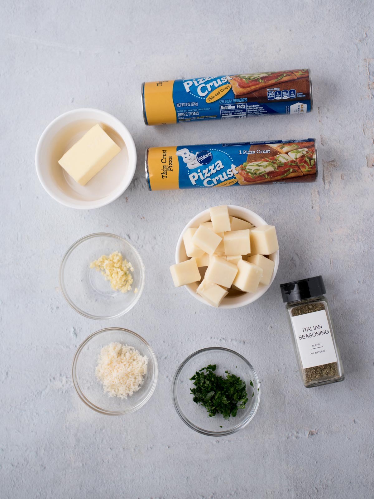 Ingredients on a light surface: two cans of pizza crust, a bowl of butter, cubed cheese, grated cheese, minced garlic, chopped herbs, and a jar labeled Italian Seasoning.