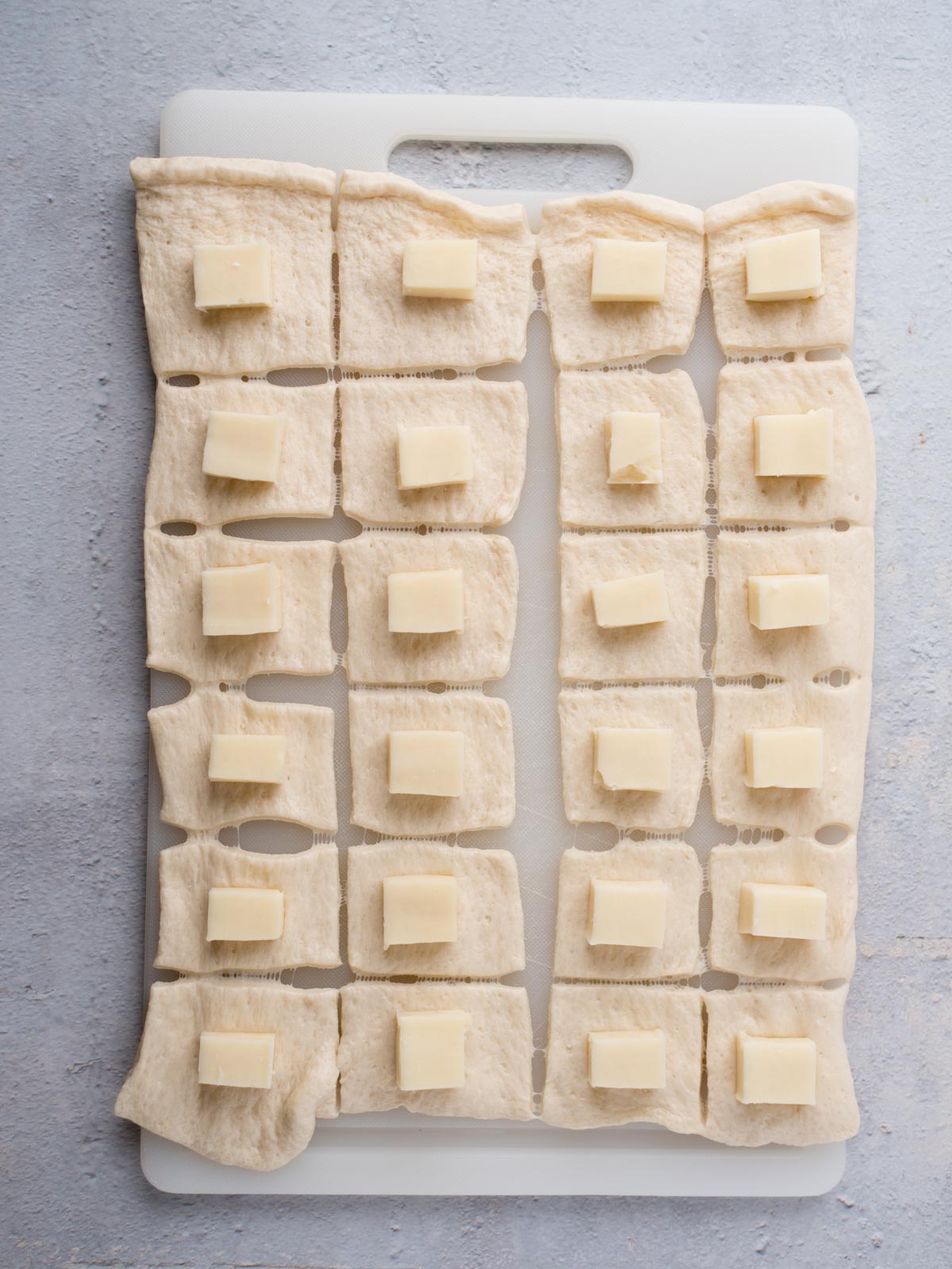 A white cutting board with rectangular dough pieces, each topped with a small square of cheese, arranged in a grid pattern on a light gray surface.