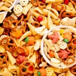 A tray and bowl filled with Halloween snack mix, including pretzels, candy corn, pumpkin candies, Chex cereal, bugle chips, and colorful chocolate candies. The image features the text, Halloween snack mix, sweetlysplendid.com.