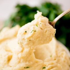 A close-up of a spoonful of creamy slow cooker mashed potatoes garnished with chopped herbs, with more mashed potatoes and green herbs blurred in the background.
