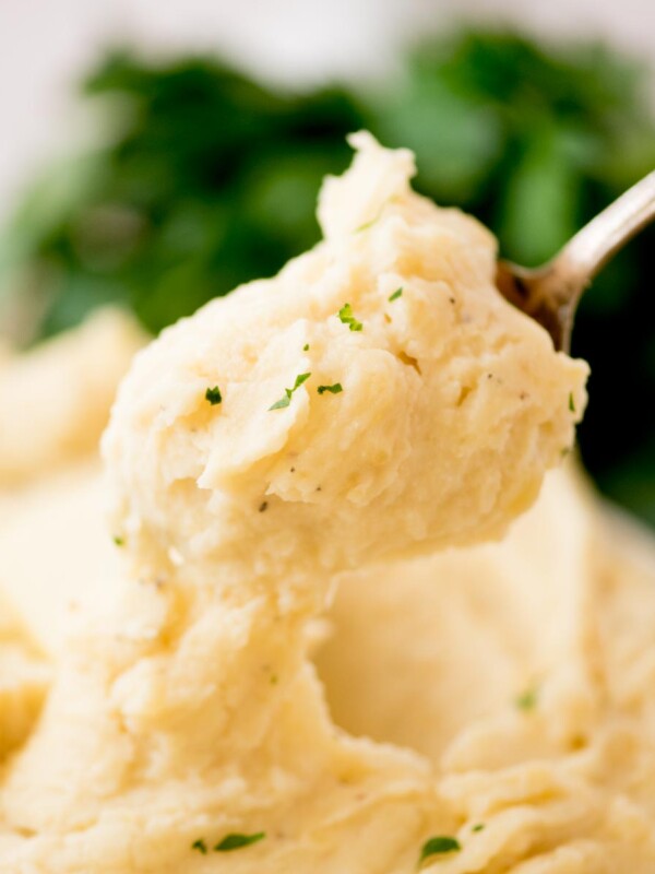 A close-up of a spoonful of creamy slow cooker mashed potatoes garnished with chopped herbs, with more mashed potatoes and green herbs blurred in the background.