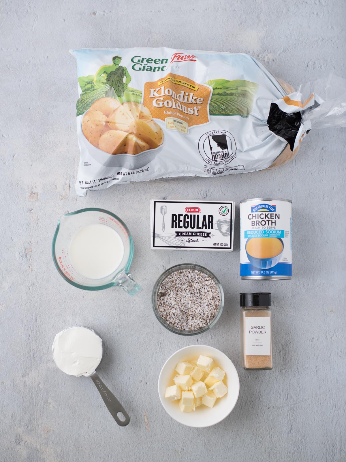 Ingredients for crockpot mashed potatoes laid out on a light surface: a bag of Klondike Goldust potatoes, milk in a measuring cup, cream cheese, chicken broth, a bowl of cubed butter, garlic powder, and a cup of sour cream.
