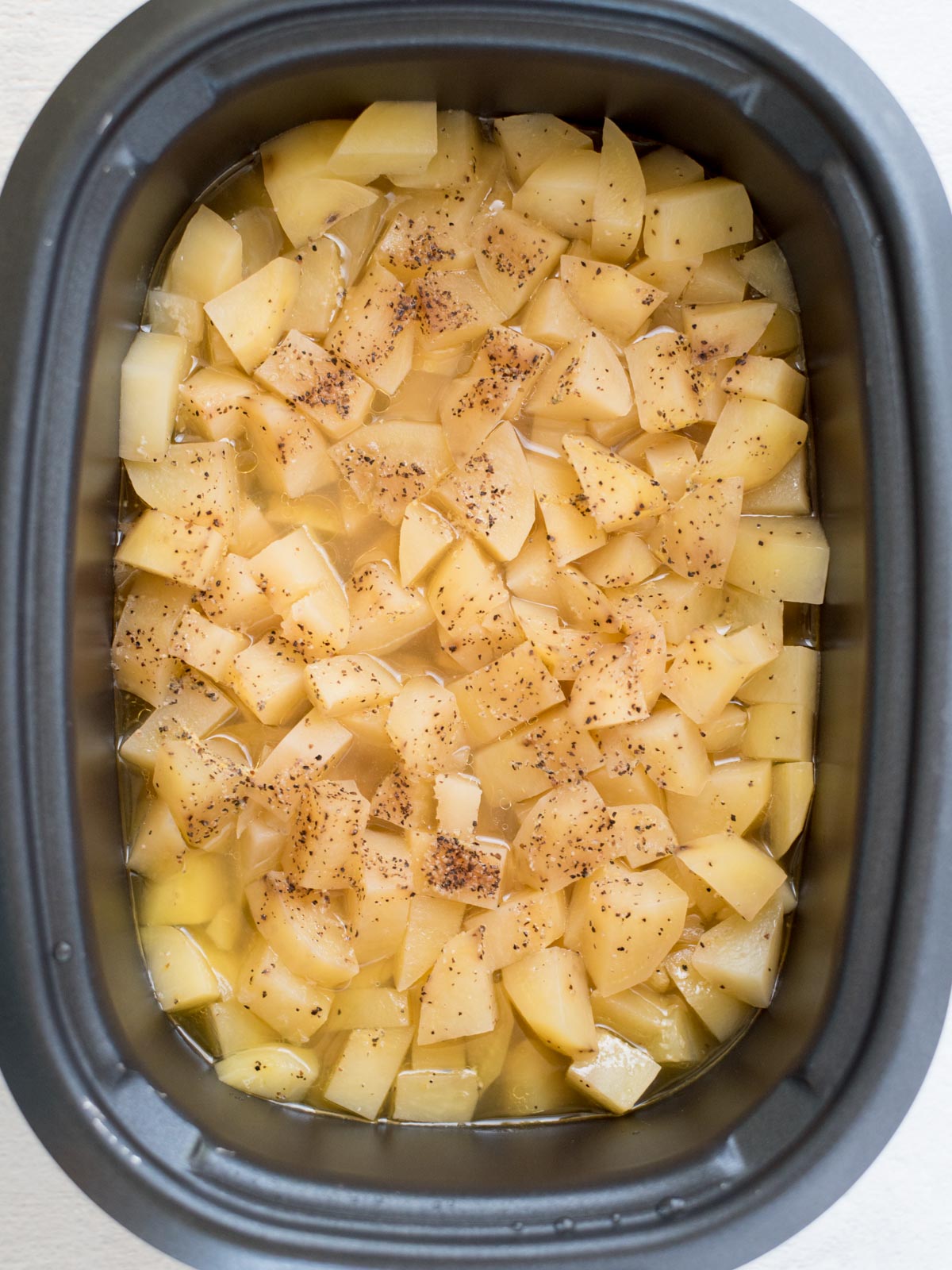 Diced potatoes seasoned with black pepper and cooking liquid inside a slow cooker, perfect for making creamy slow cooker mashed potatoes, viewed from above.
