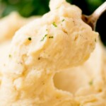 A close-up of creamy mashed potatoes being scooped with a spoon, garnished with chopped herbs. Text at the top reads “slow cooker mashed potatoes” with the website sweetlysplendid.com below.
