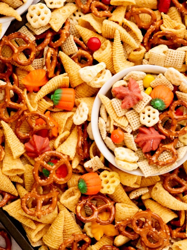 A close-up of snack mix with pretzels, Bugles, cereal pieces, candy pumpkins, candy corn, and colorful chocolate candies, with a small white bowl filled with the mix in the center.
