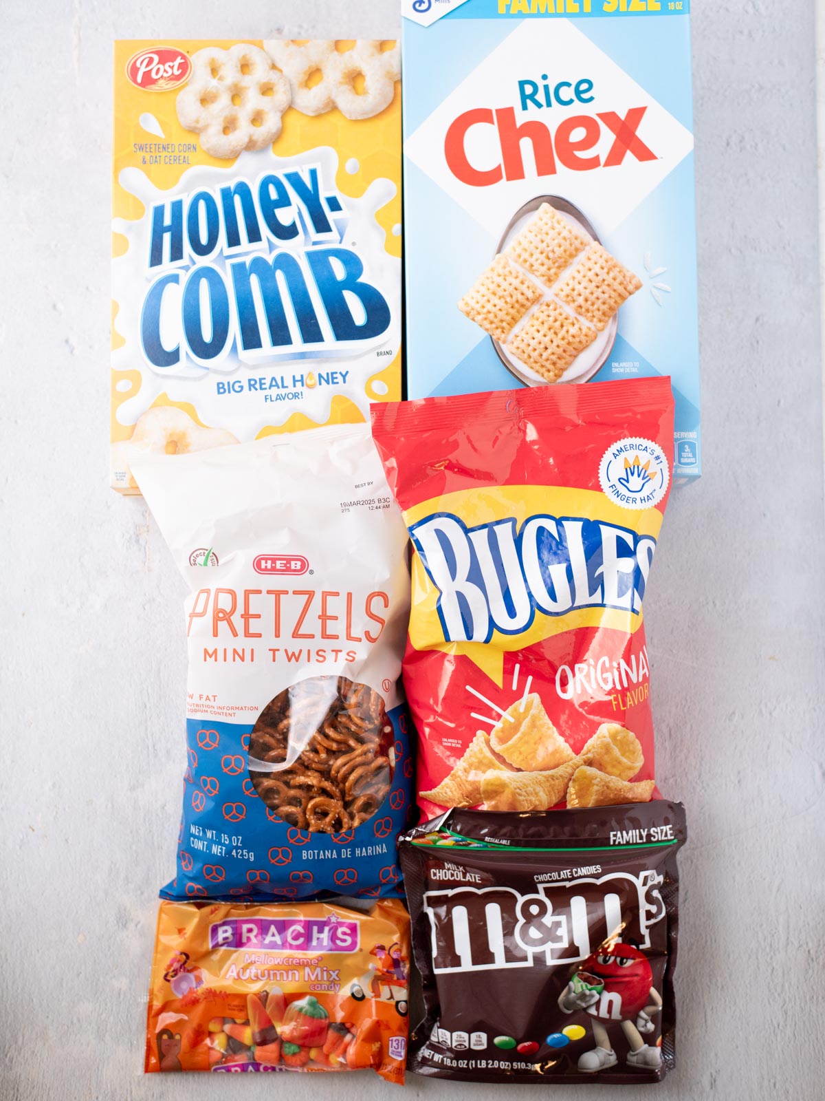 Six snack packages—Honeycomb cereal, Rice Chex, H-E-B pretzels, Bugles, Brach’s Autumn Mix candy, and M&M’s—arranged on a light surface in two rows.