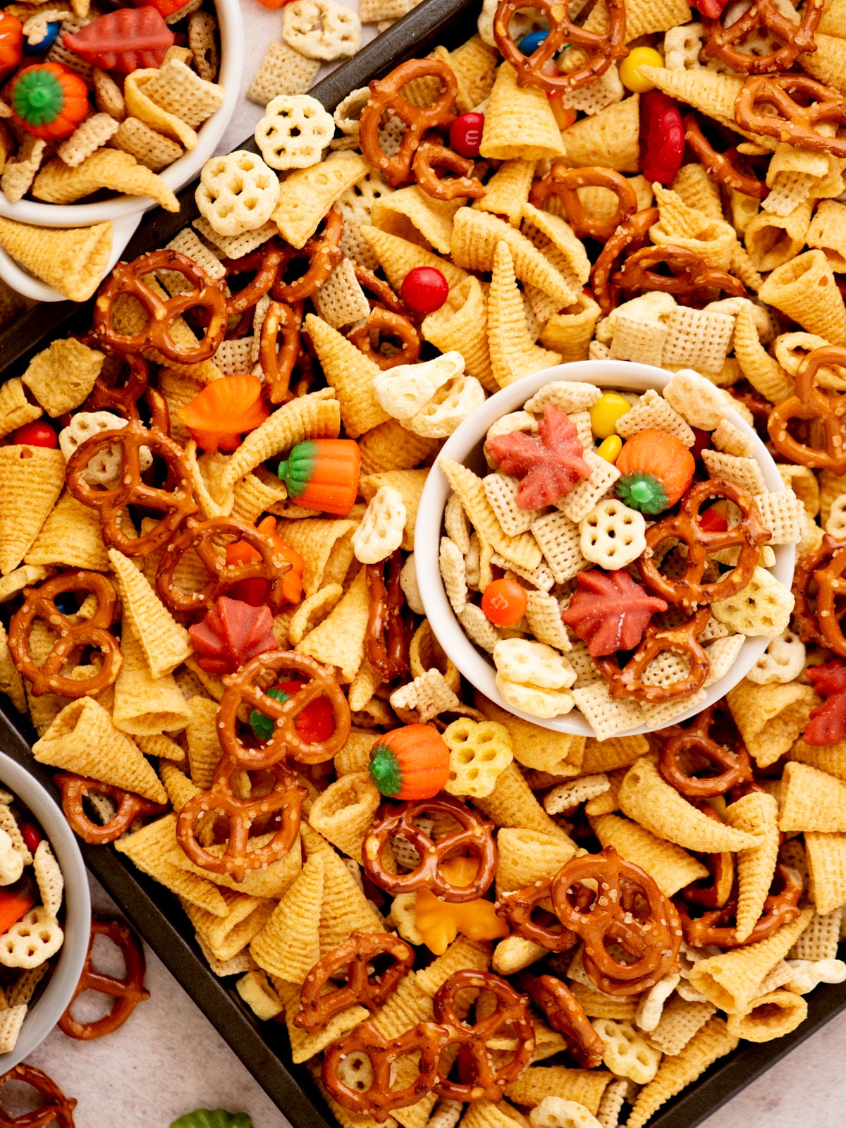A festive snack mix featuring pretzels, Bugles, Chex cereal, candy pumpkins, and colorful candy-coated chocolates, displayed in bowls and scattered on a tray.