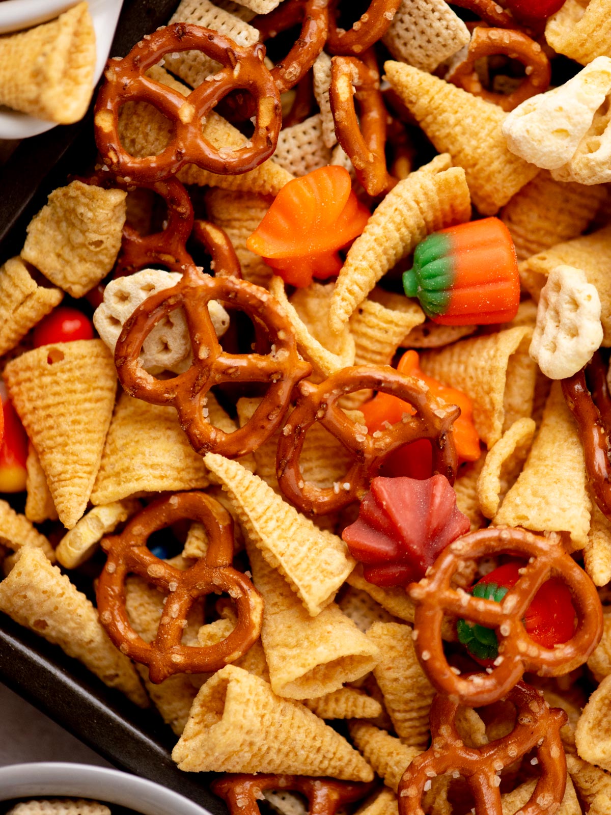 A close-up of a snack mix featuring pretzels, bugle chips, Chex cereal, and candy pumpkins and leaves in fall colors, all piled together in a tray.