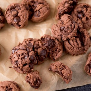 A close-up of several chocolate chocolate-chip cookies on a sheet of brown parchment paper, showing gooey chocolate chips and a soft, crumbly texture, with a few cookies broken in half.