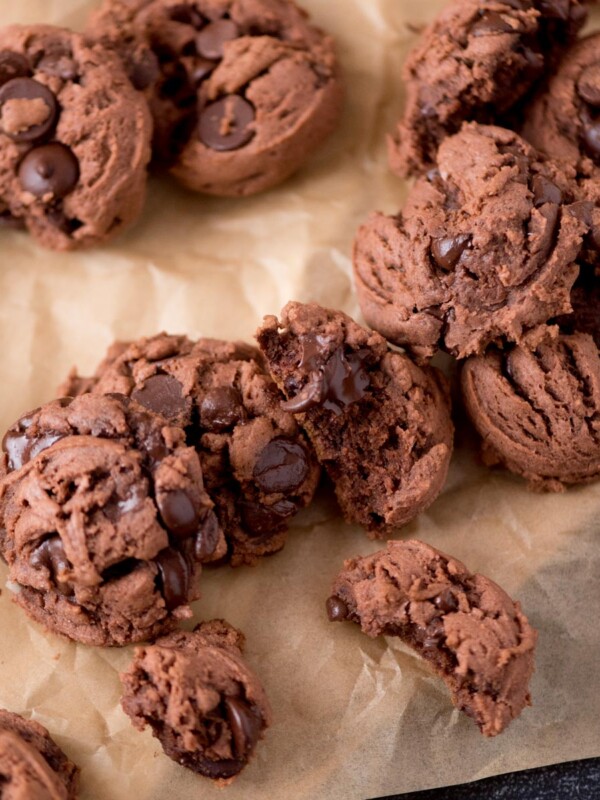 A close-up of several chocolate chocolate-chip cookies on a sheet of brown parchment paper, showing gooey chocolate chips and a soft, crumbly texture, with a few cookies broken in half.