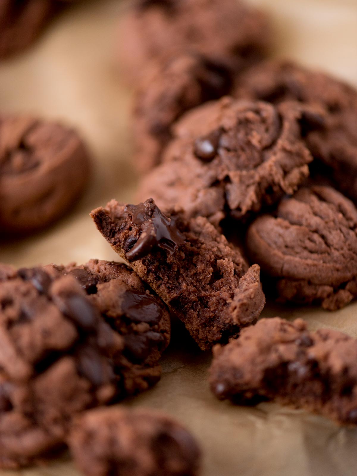 A close-up of several chocolate cookies with chocolate chips, some broken in half, resting on a sheet of brown parchment paper.