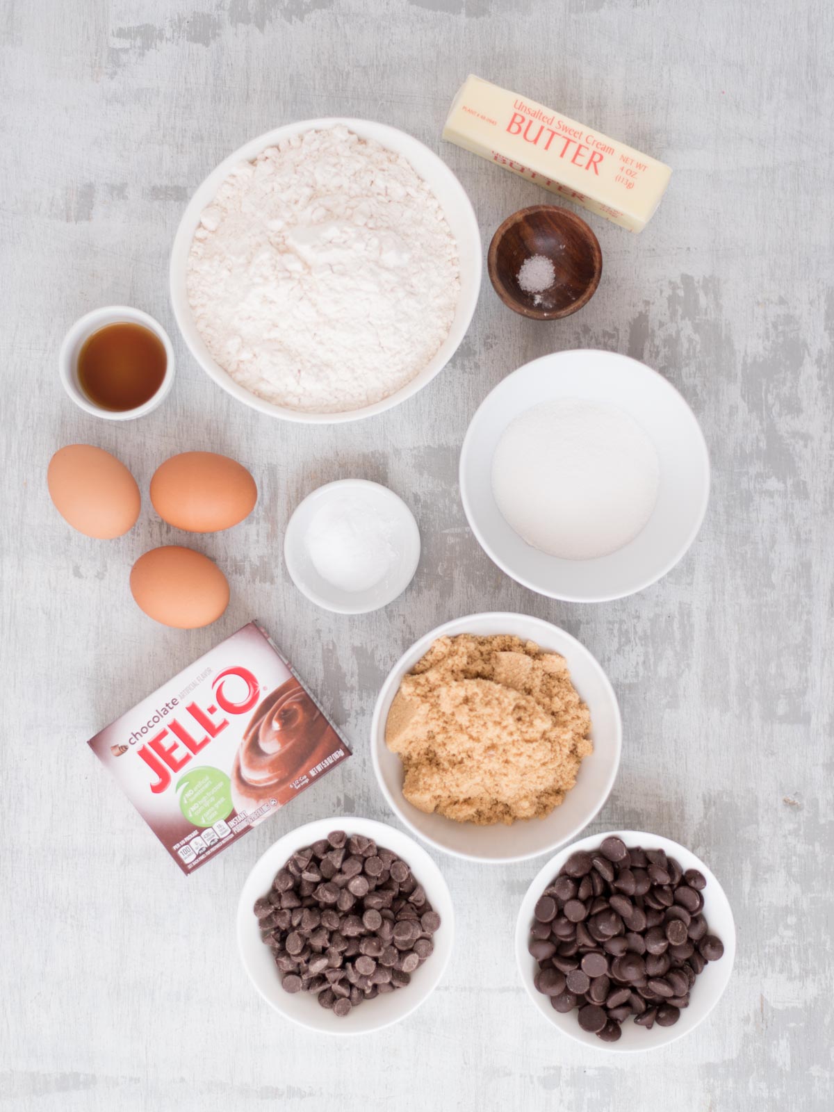 A flat lay of baking ingredients includes flour, eggs, butter, vanilla extract, salt, granulated sugar, brown sugar, baking soda, chocolate pudding mix, and two bowls of chocolate chips on a light surface.