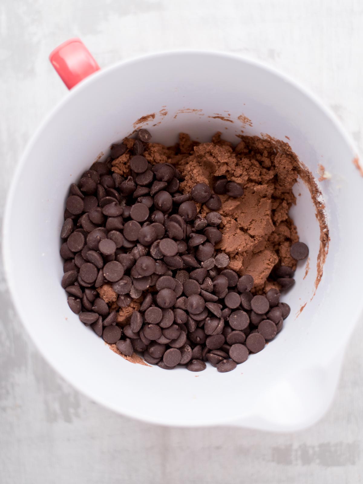 A white mixing bowl with a red handle contains chocolate cookie dough and a generous amount of chocolate chips on a light-colored surface.