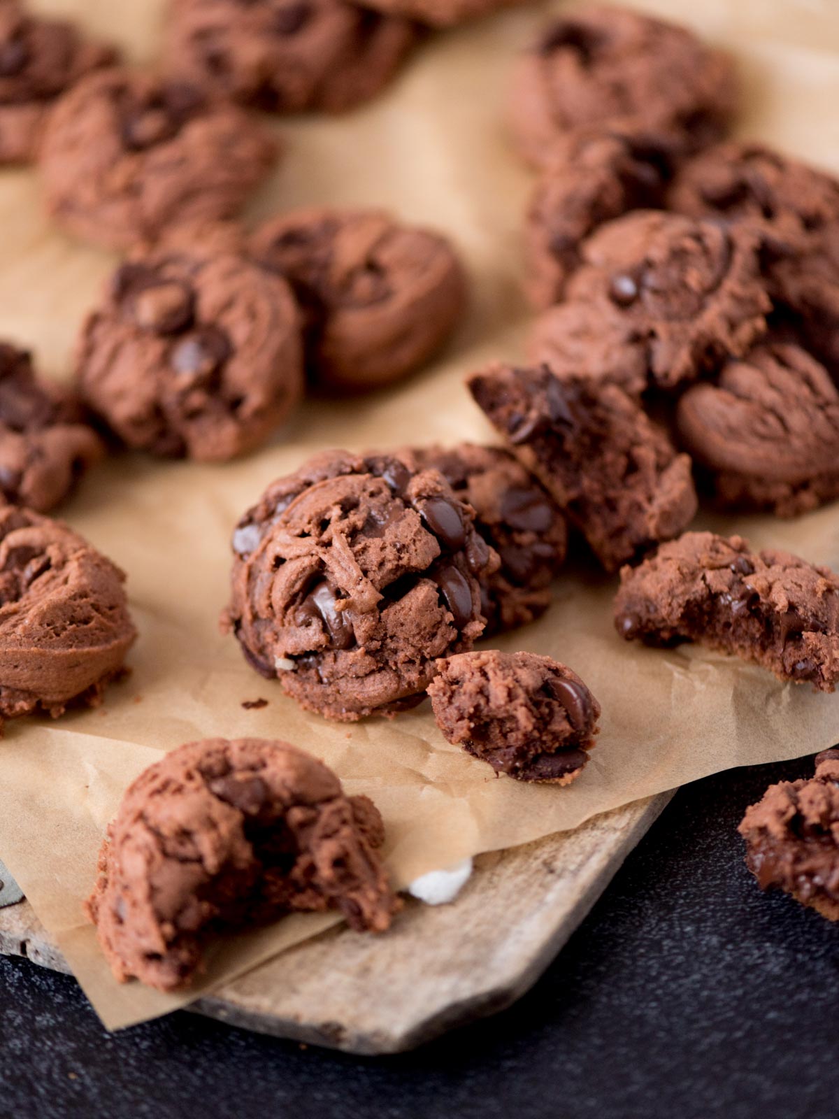 A close-up of several chocolate cookies, some whole and some broken, resting on brown parchment paper atop a wooden board. The cookies are studded with chocolate chips.