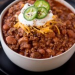 A bowl of ground beef chili with beans, topped with shredded cheese, sour cream, and sliced jalapeños, is placed on a black plate with fresh cilantro nearby. A sign above reads, ground beef chili with beans.