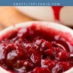 A white bowl filled with thick, chunky cranberry sauce sits on a surface, with more cranberry sauce visible in the background. The image includes the text cranberry sauce and sweetlysplendid.com at the top.