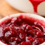 A white bowl filled with homemade cranberry sauce sits on a table, with a dish of cranberry sauce in the background. A label above the image reads homemade cranberry sauce, sweetlysplendid.com.