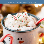 A festive mug with a snowman face is filled with hot cocoa, topped with whipped cream, holiday sprinkles, mini marshmallows, and a candy cane. Warm holiday lights and decorations are blurred in the background.