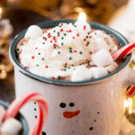 A festive mug decorated with a snowman face is filled with hot chocolate, whipped cream, mini marshmallows, red and green sprinkles, and a candy cane. Holiday lights and decorations are blurred in the background.