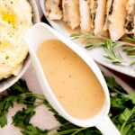 A white gravy boat filled with turkey gravy sits on a table next to mashed potatoes, sliced roast meat, rosemary sprigs, and fresh parsley.