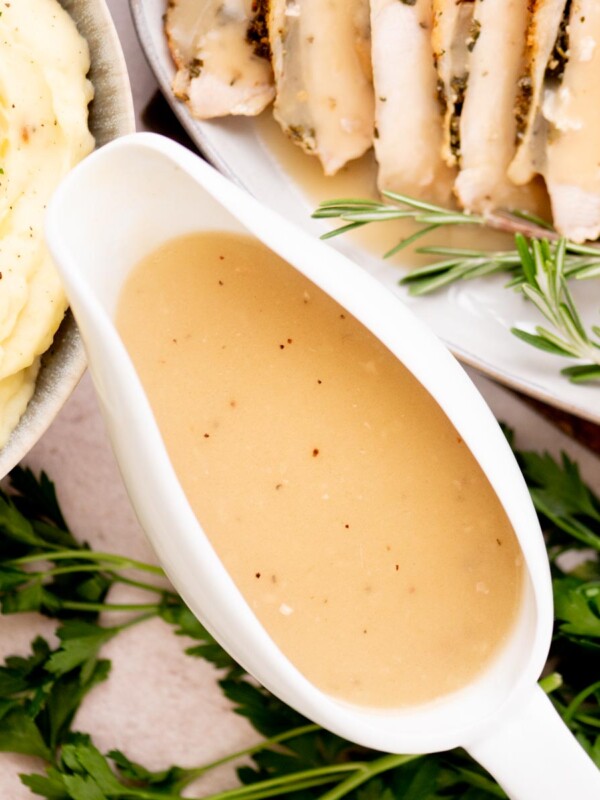A white gravy boat filled with turkey gravy sits on a table next to mashed potatoes, sliced roast meat, rosemary sprigs, and fresh parsley.