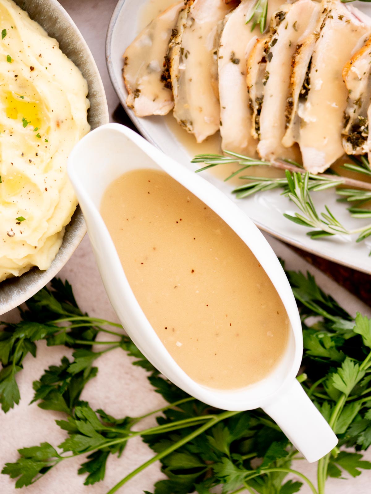 A white gravy boat filled with creamy brown thanksgiving gravy sits on a table next to a bowl of mashed potatoes and a plate of sliced roast chicken garnished with fresh rosemary and parsley.