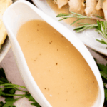 A white gravy boat filled with golden turkey gravy, surrounded by fresh herbs and a plate of sliced turkey, with a sign reading turkey gravy from drippings at the top.