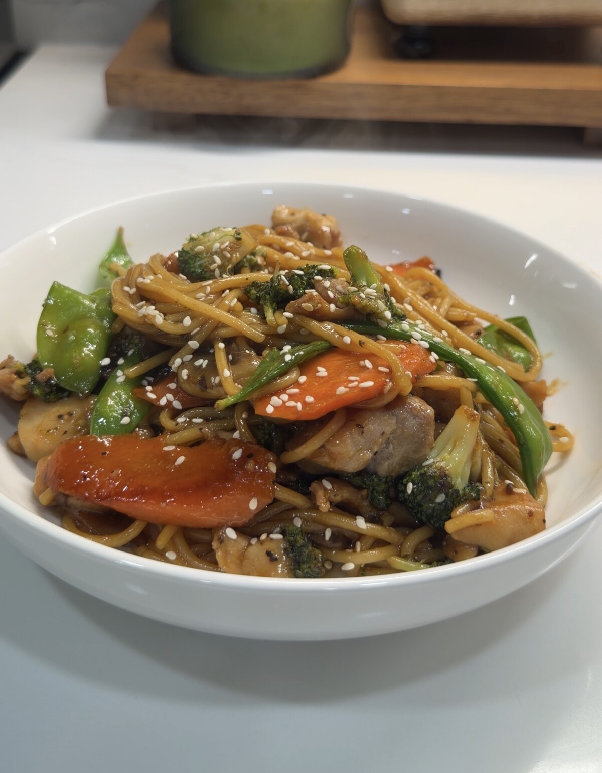 A white bowl filled with Chicken Teriyaki noodles, chicken, broccoli, carrots, snap peas, and mushrooms, garnished with sesame seeds, on a white countertop.