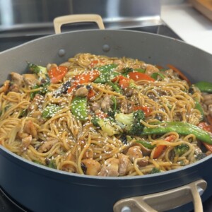 A large pan filled with Chicken Teriyaki noodles, broccoli, snap peas, carrots, and red bell peppers, topped with sesame seeds, sits on a stove.