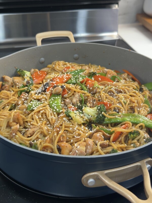 A large pan filled with Chicken Teriyaki noodles, broccoli, snap peas, carrots, and red bell peppers, topped with sesame seeds, sits on a stove.
