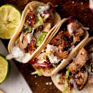 Three soft tacos filled with grilled chicken, shredded lettuce, cabbage, cheese, and creamy sauce are arranged on a wooden board with halved limes beside them.
