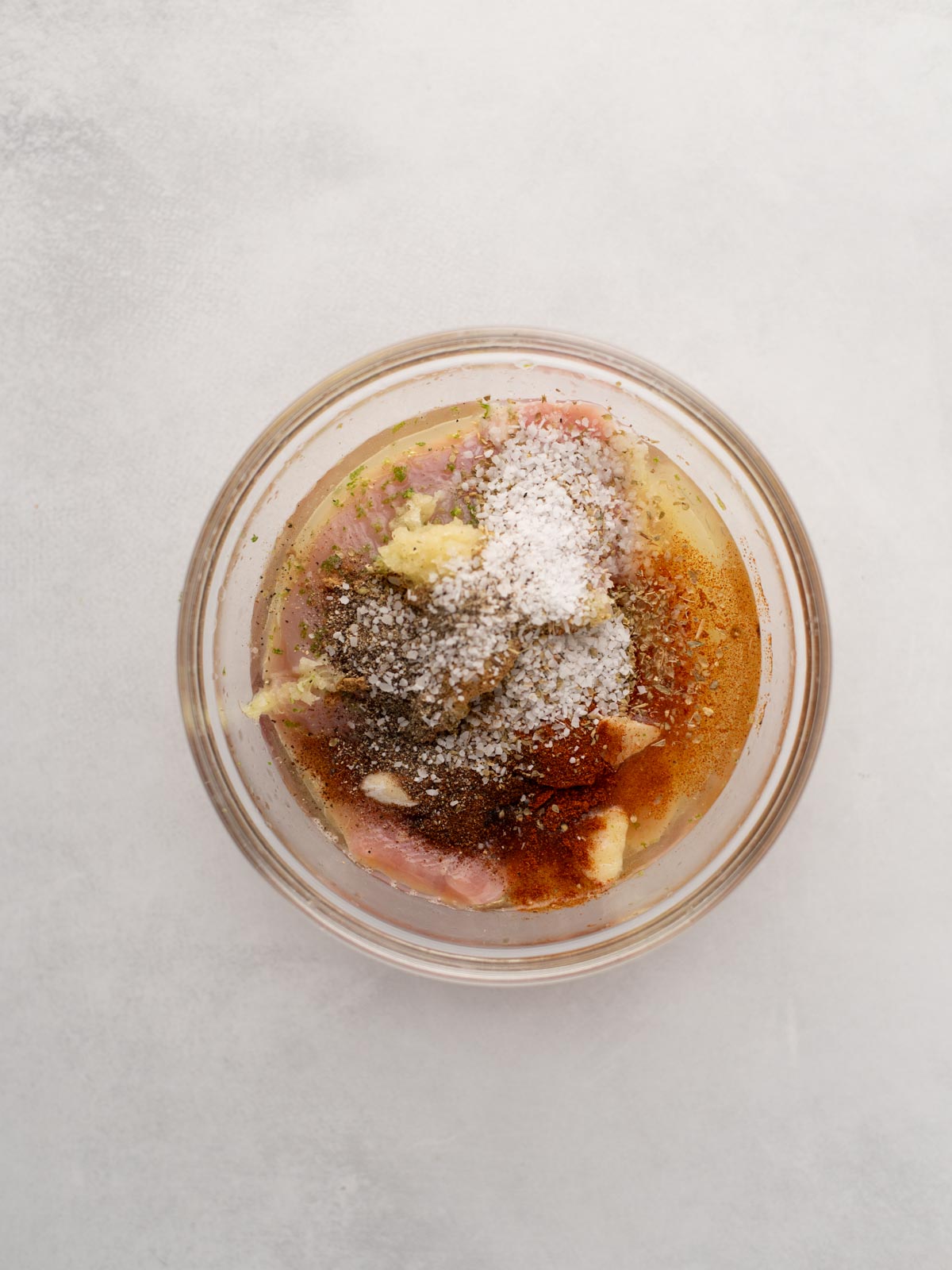 A glass bowl containing raw chicken pieces, garlic cloves, various spices, and salt, all ready to be mixed for marinating, set on a light gray surface.