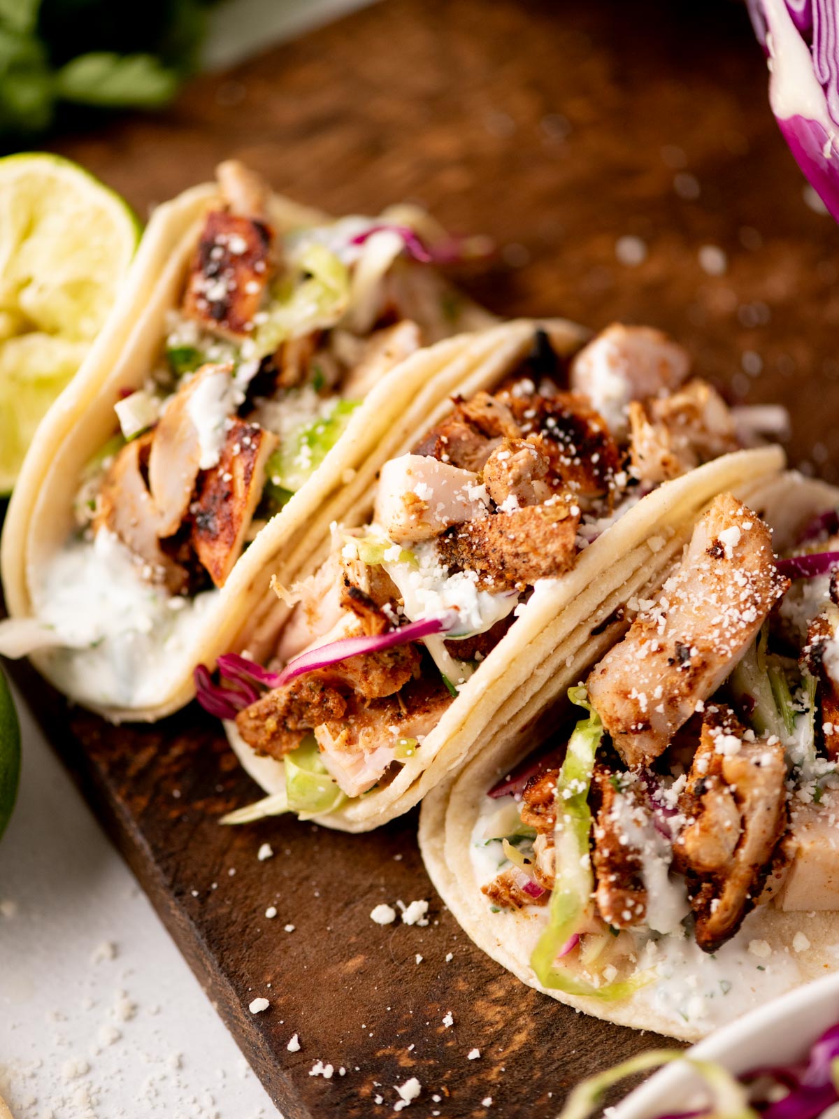 Three soft tacos filled with grilled chicken, shredded lettuce, red cabbage, creamy sauce, and crumbled cheese are arranged on a wooden board. A halved lime sits nearby.