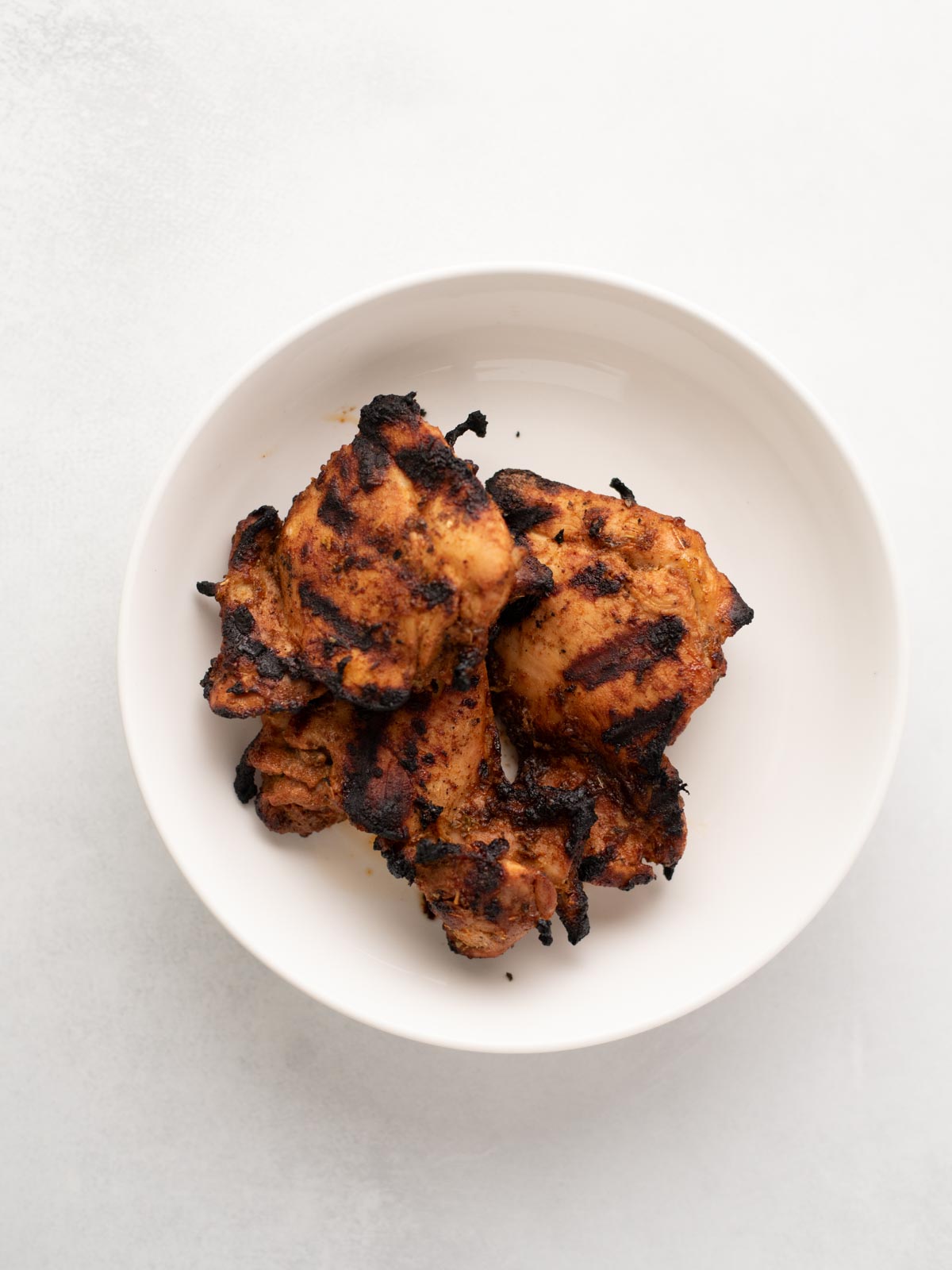 A white bowl containing several pieces of grilled, charred chicken with visible grill marks, set against a light background.