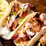 Three chicken street tacos filled with grilled chicken, shredded lettuce, and creamy sauce sit on a wooden board. A wedge of lime is beside them. A text banner at the top reads “chicken street tacos.”.