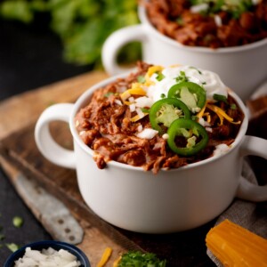 A white bowl of chili topped with sliced jalapenos, shredded cheese, chopped onions, and a dollop of sour cream, sits on a wooden board with fresh herbs and a block of cheddar cheese nearby.
