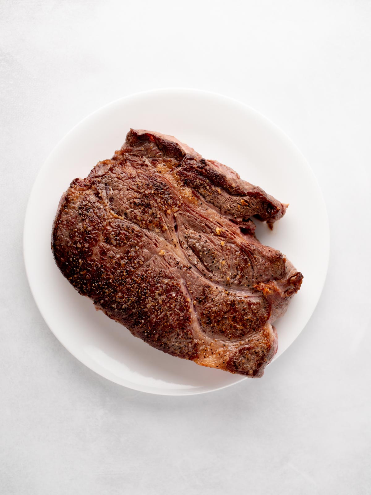 A cooked, seasoned steak sits on a white plate against a light background. The steak has a browned, slightly charred exterior with visible spices.