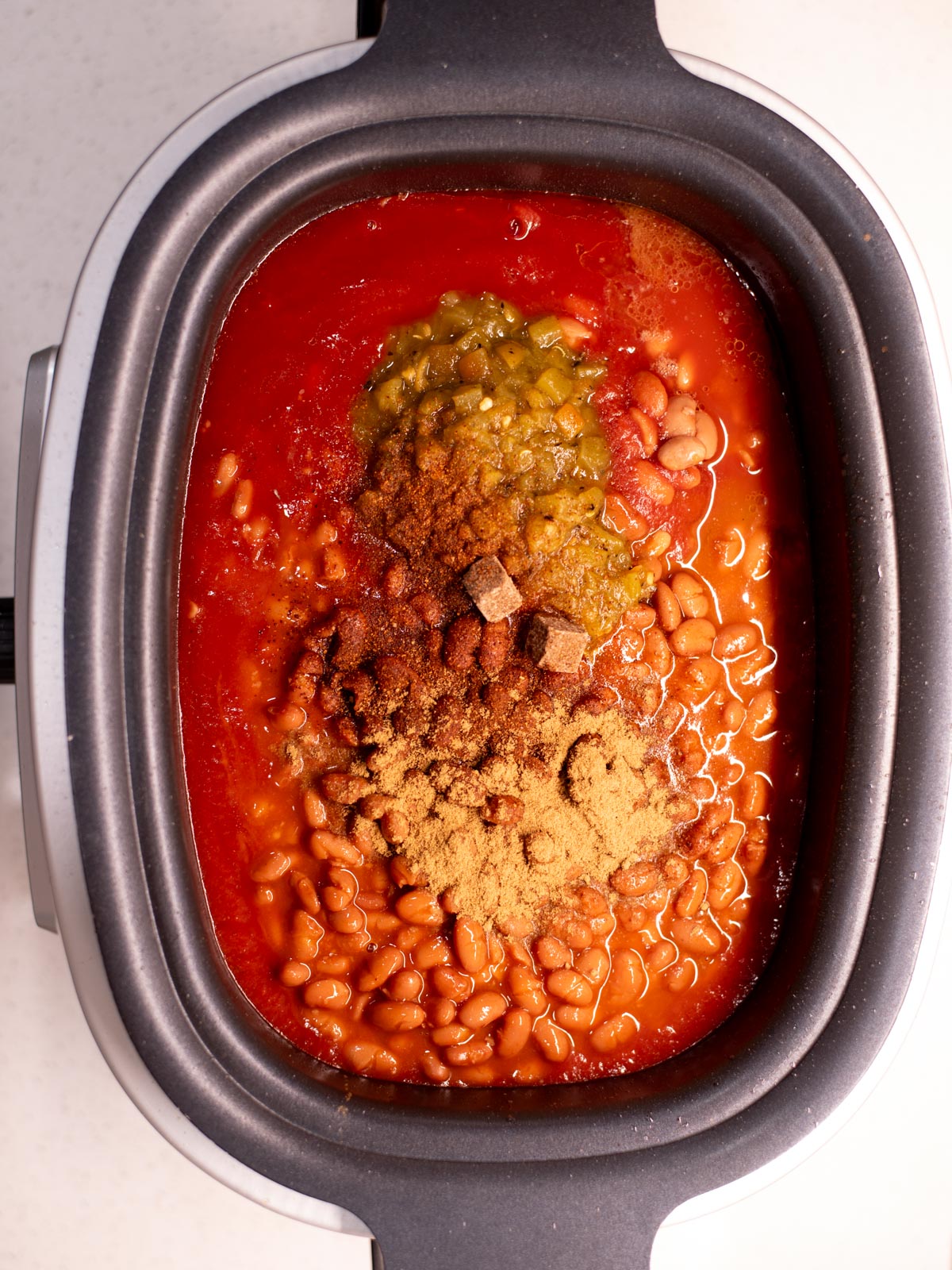 Overhead view of a slow cooker filled with baked beans, tomato sauce, chopped green chilies, and various spices, not yet mixed together.