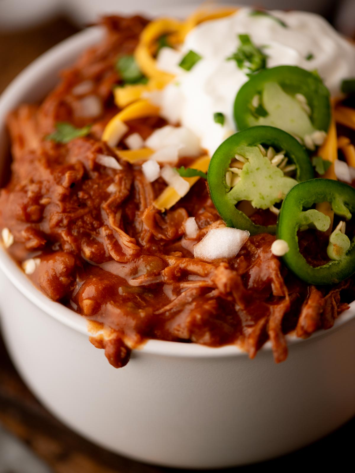 A bowl of chili topped with sliced jalapeños, shredded cheddar cheese, sour cream, chopped onions, and fresh cilantro. The chili appears thick and hearty with shredded meat.