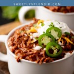 A white mug filled with chili, topped with sour cream, shredded cheese, jalapeño slices, and chopped cilantro, sits on a wooden board. Text above reads “chuck roast chili” and “sweetlysplendid.com.”.