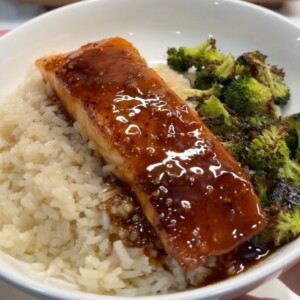 A bowl filled with white rice, glazed salmon topped with a shiny sauce, and roasted broccoli florets on the side.