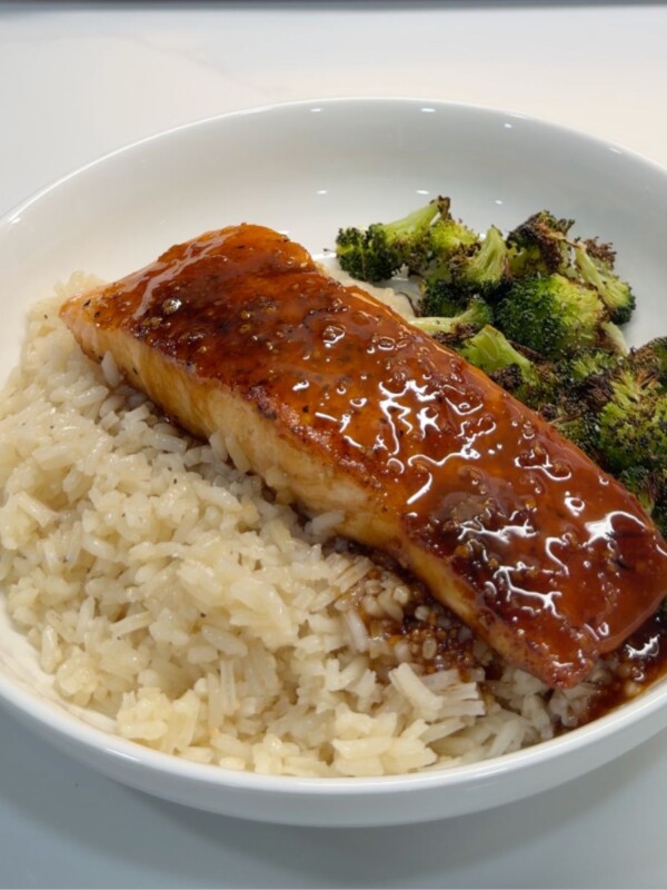 A white bowl filled with white rice, glazed salmon filet topped with sauce, and roasted broccoli, all placed on a white surface.