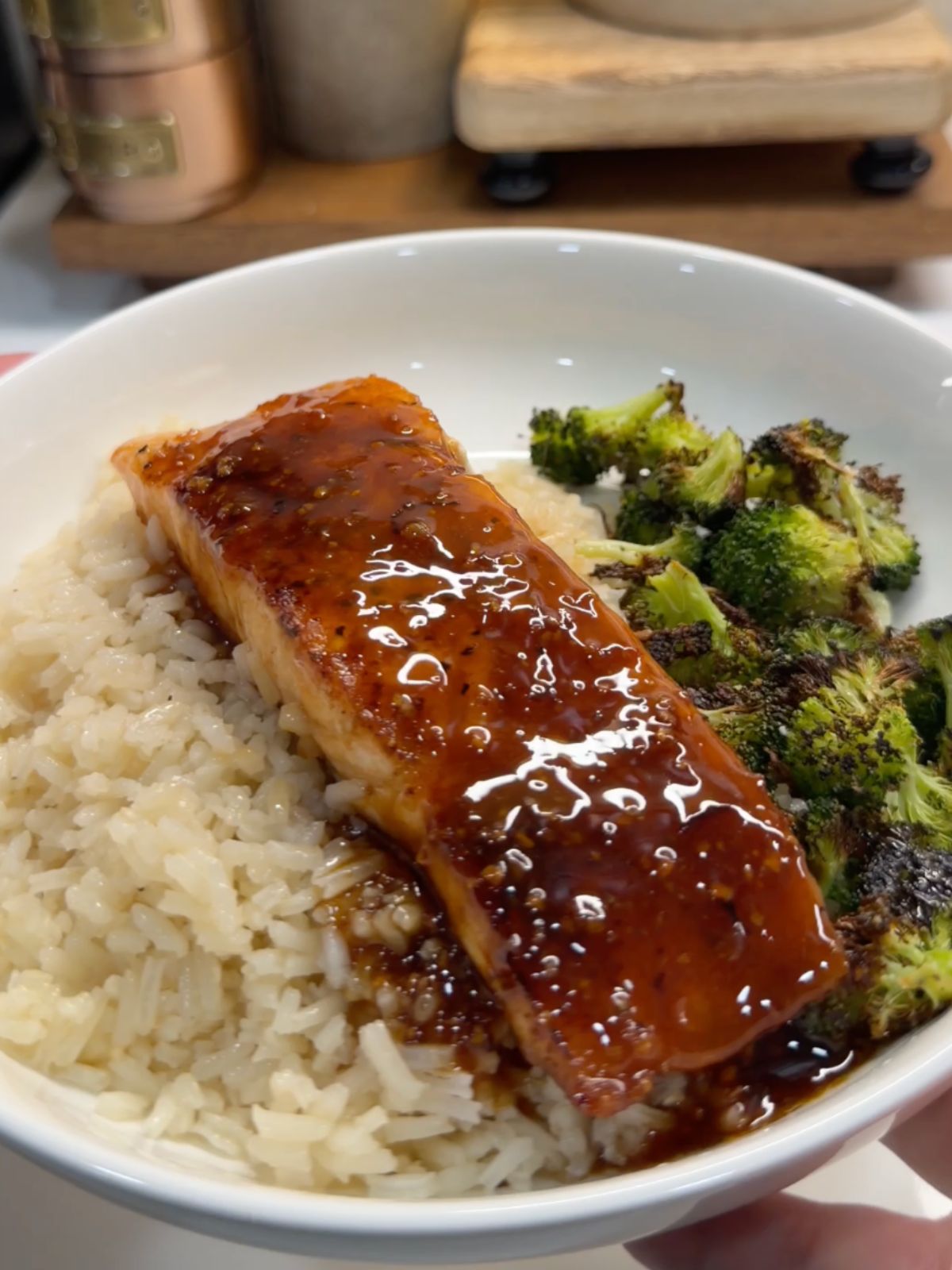 A bowl of white rice topped with a glazed salmon fillet and served with roasted broccoli florets. The glaze on the salmon is shiny and caramelized.